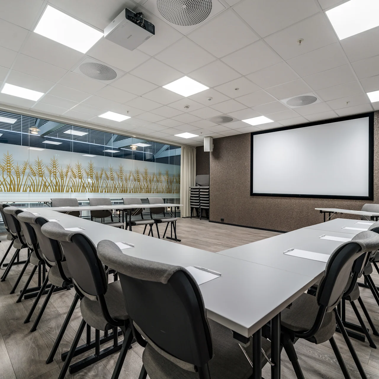 Conference room with chairs arranged in a U-shape around tables, facing a large projection screen. The room has wood flooring and frosted glass windows with wheat designs.