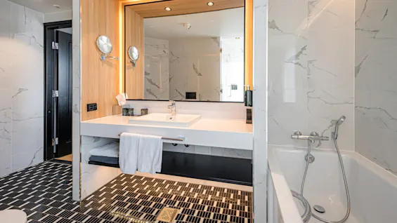 A modern bathroom features a white sink and countertop with a large mirror, surrounded by sleek lighting and marble walls. A bathtub with a handheld showerhead is to the right.