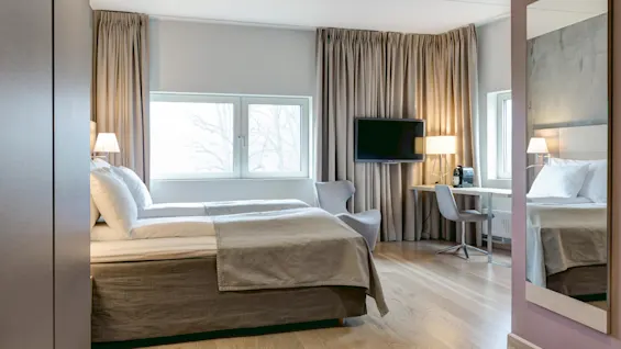 A neatly made bed stands in a modern hotel room, accompanied by a television on the wall, a desk, chair, and window with curtained view, set on wooden flooring.