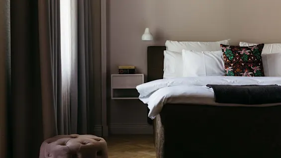 Seng med hvite puter og en fargerik pute, i et koselig rom med gardiner og et lite sidebord med bøker. Varm belysning forsterker den fredelige atmosfæren.