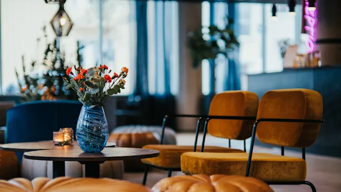 A blue glass vase with red flowers sits on a table in a stylish lounge with mustard-yellow chairs, leather poufs, and soft lighting. Neon sign partially visible.