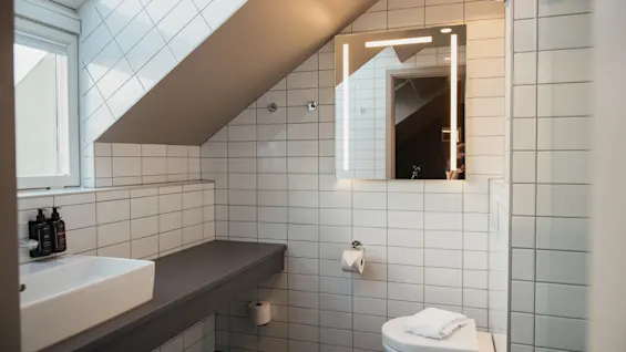 A bright, modern bathroom with white tiles, a sloped ceiling, and an illuminated mirror, creating a clean and functional space.