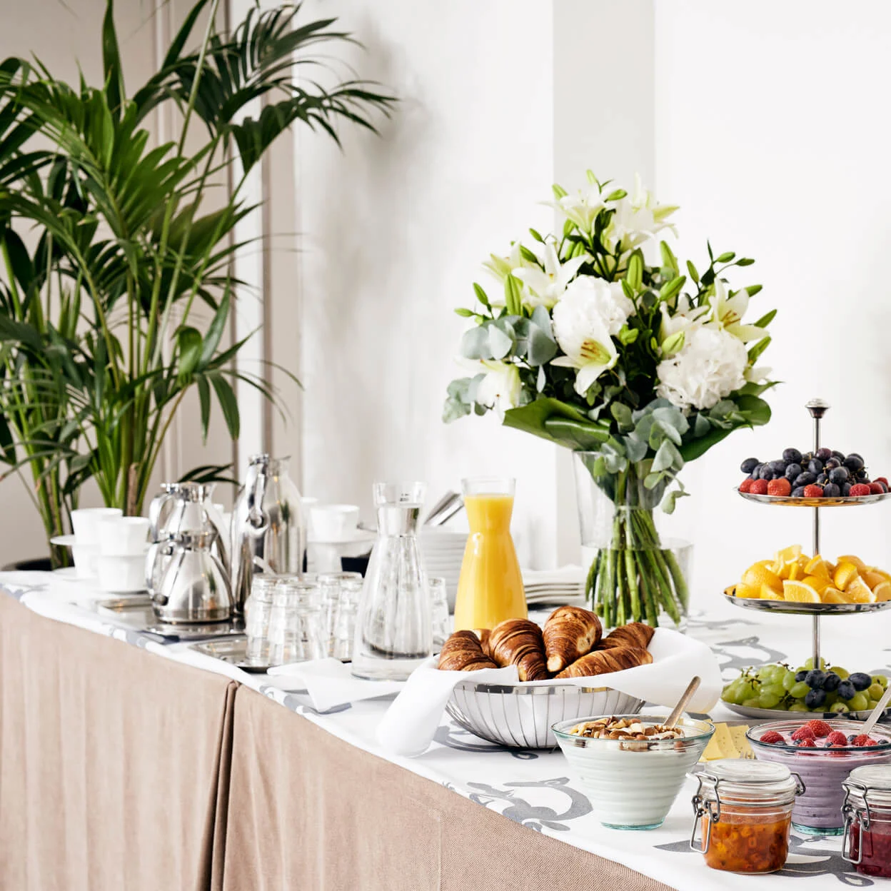 Breakfast buffet with croissants, juices, fruits, and yogurts on a decorated table, surrounded by green plants and white flowers, creating an elegant dining atmosphere.