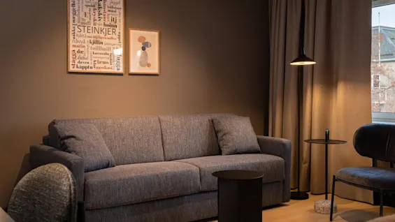 A cozy hotel room with a grey sofa, a floor lamp, and a side table. Wall art includes text like STEINKJER.