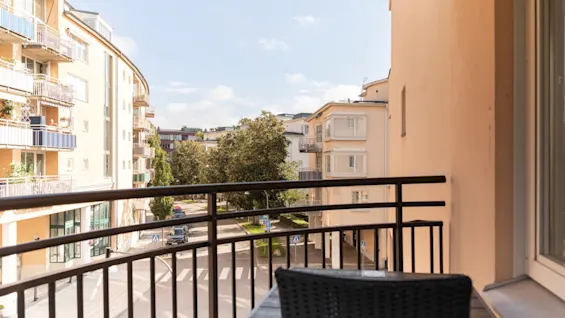 Utsikt från en hotellbalkong med utsikt över en gata med byggnader och träd under en klar himmel. Ett bord och en stol finns på balkongen.