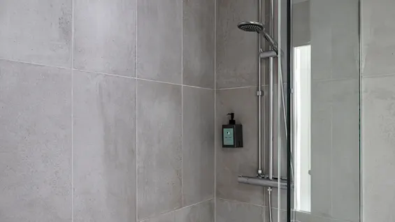 En moderne dusj med stilige grå fliser og dør av glass uten ramme. Et regndusjhode er montert over et mindre håndholdt alternativ, med en elegant såpedispenser på veggen. Mykt lys siver inn.