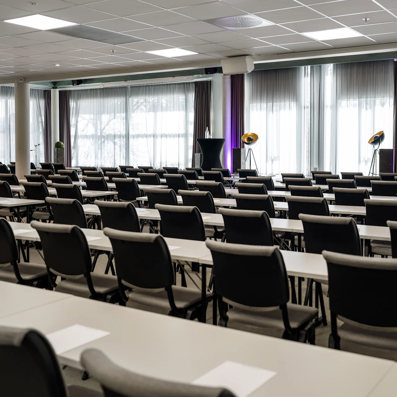 Rows of gray chairs and desks face a podium in an empty conference room. Large windows with sheer curtains let in light, with purple uplights highlighting the walls.