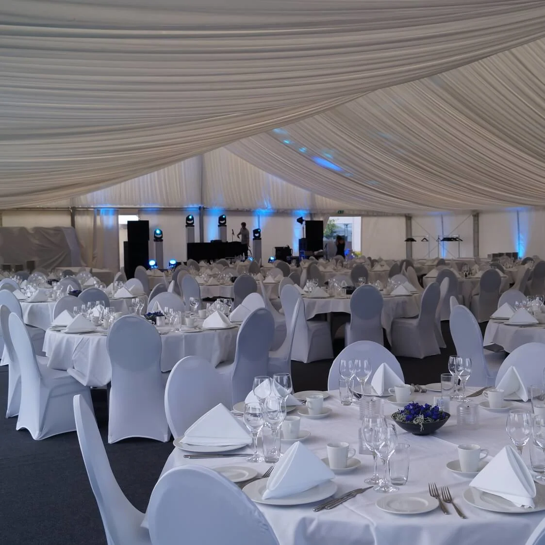 Round tables with white tablecloths set with glassware and napkins, surrounded by chairs, inside a spacious, draped marquee, ready for an event.