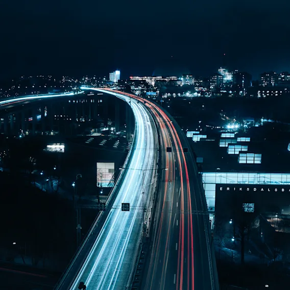 Street lights create streaks across a highway at night, with city buildings illuminated in the background. Text: "ERIKSDALSBADET".