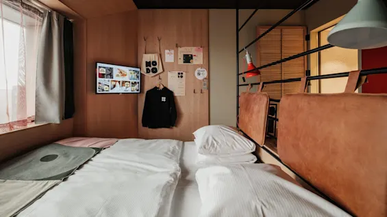Bed with white linens sits in a cozy room. Nearby, a wall displays a black garment and pegboard with various items, including a bag labeled "NORD," under a mounted screen showing images.