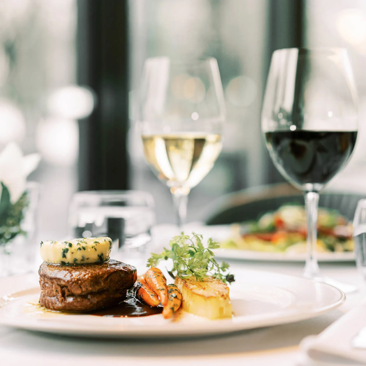 A steak with herb butter on top, side of potatoes and vegetables, with a glass of white wine, and another of red wine, elegant dining setting at Hotel Kämp in Helsinki.