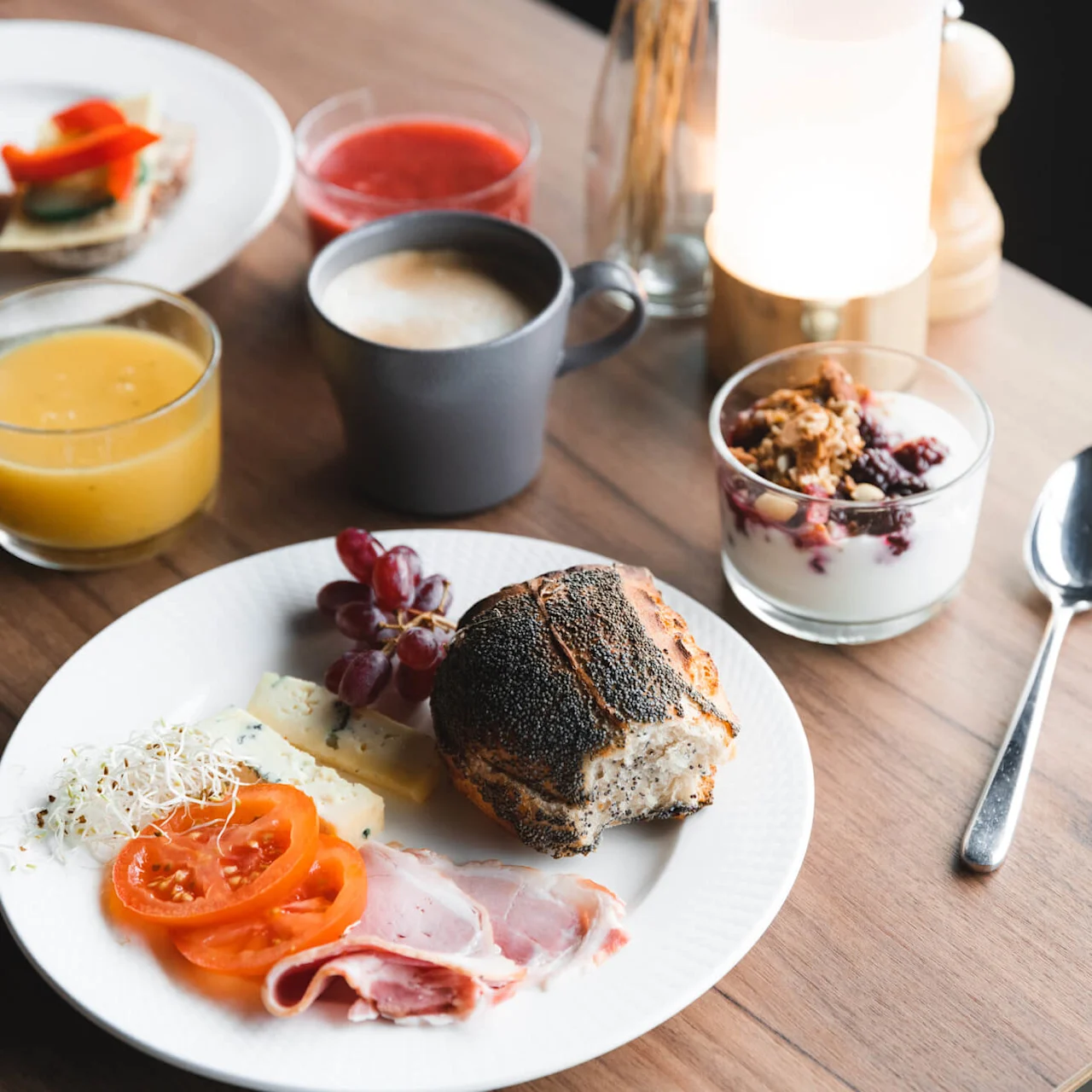 En frukosttallrik med bröd, kallskuret, ost och vindruvor står på ett träbord tillsammans med en kopp kaffe, juice, yoghurt med müsli och en sked.