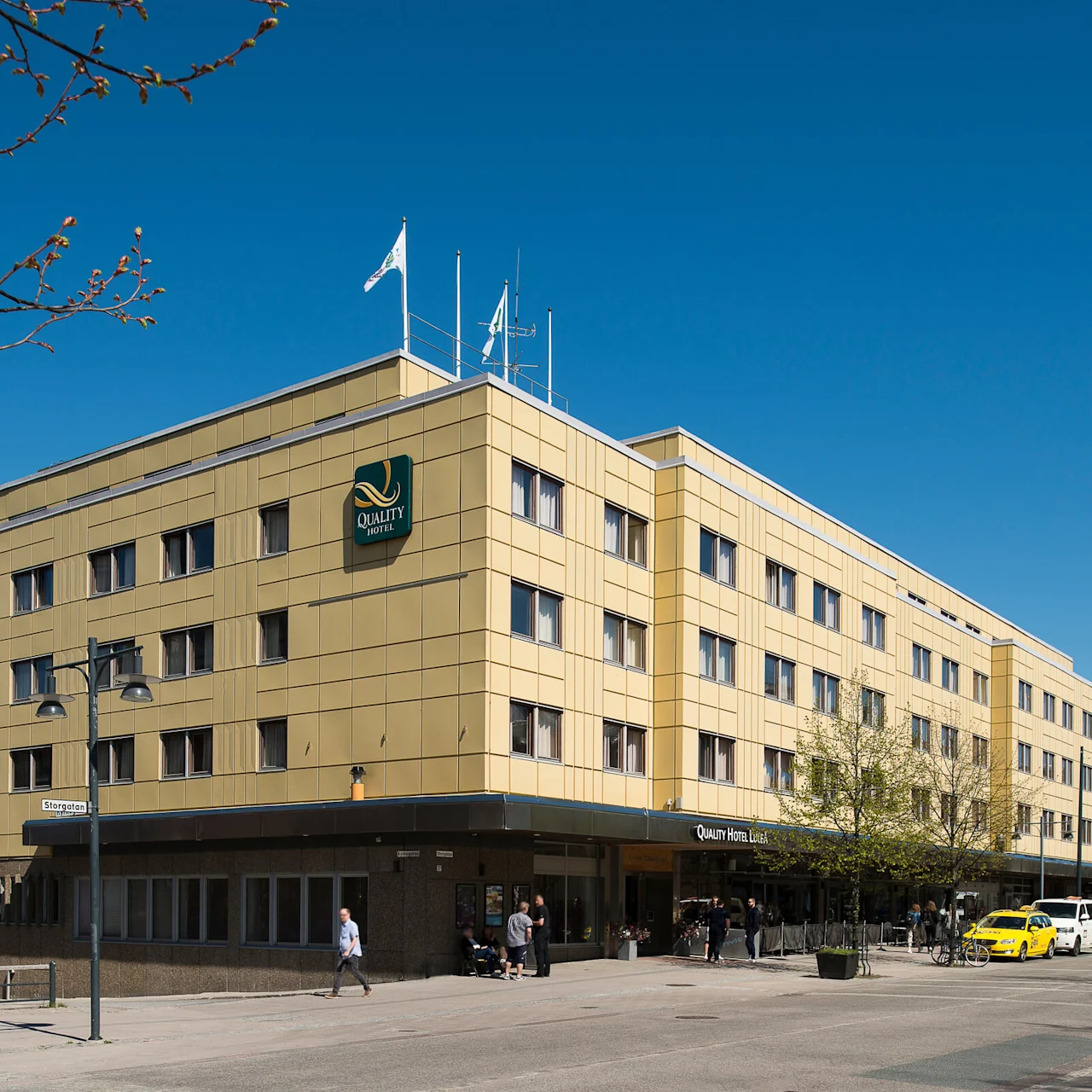 En stor gul byggnad med texten "Quality Hotel" och flera fönster står på ett gatuhörn. Fotgängare promenerar förbi och en gul taxi är parkerad i närheten under klar himmel.