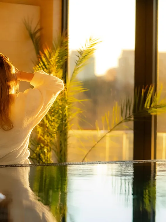 A person in a white robe stands near an indoor pool, facing a large window. Sunlight filters through, illuminating plants and casting a warm glow.