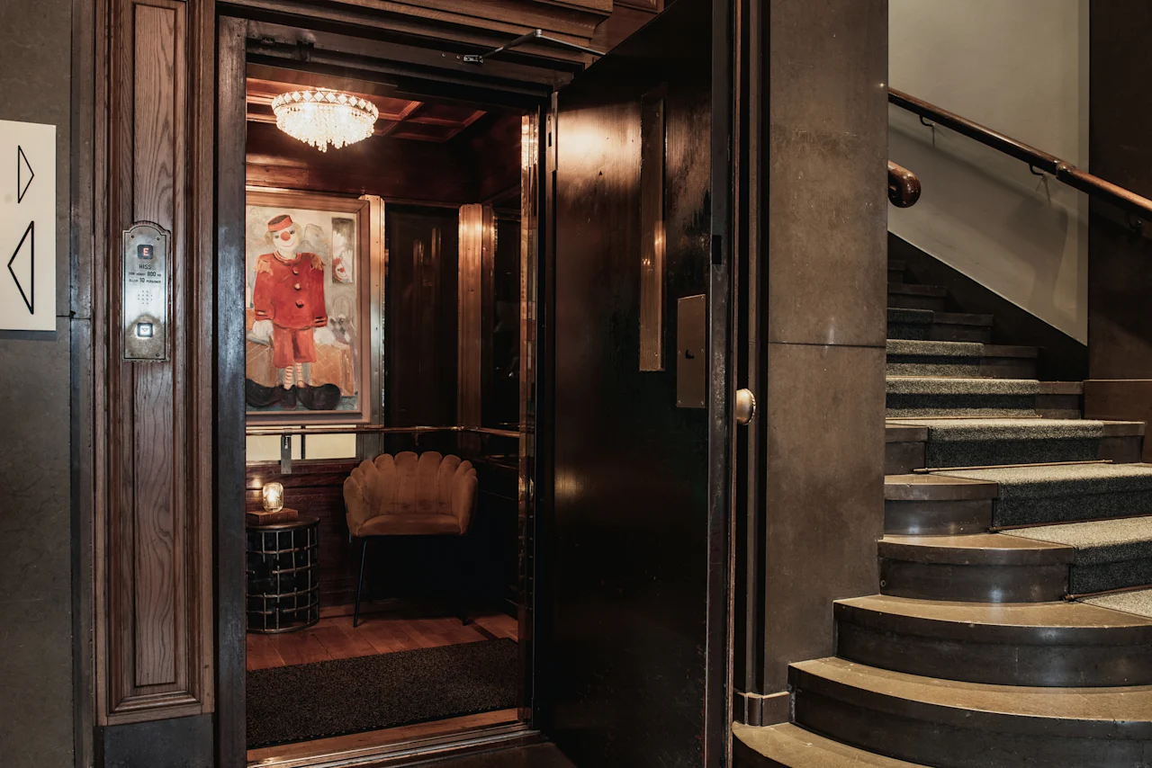 En inbjudande öppen hiss med en unik målning och en mysig stol, bredvid en storslagen trappa på Home Hotel Grand, Helsingborg.