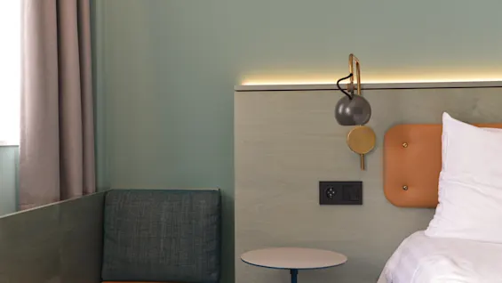 Seng med hvide puder og et orange sengegærde, ved siden af en lampe på en træpanelvæg, ved siden af en turkis pude og et lille rundt bord i et rum med dæmpede grønne vægge.