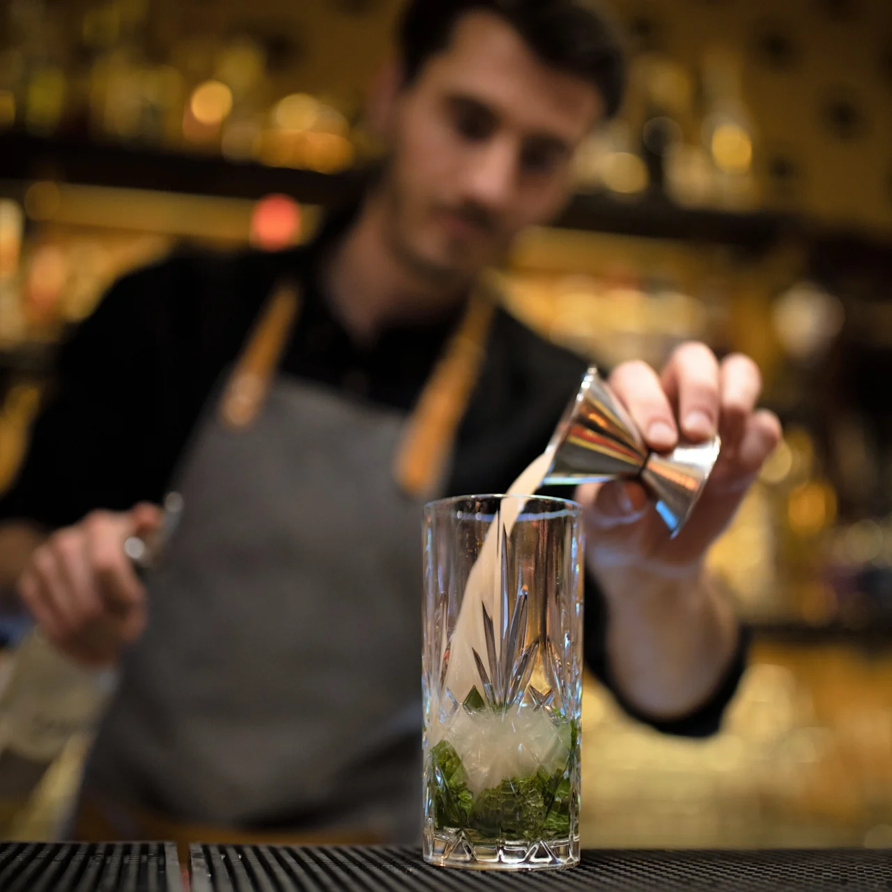 En bartender häller vätska i ett dekorerat glas med is och mynta med hjälp av en mätkopp, stående bakom en bardisk, mot en suddig bakgrund av flaskor på hyllor.