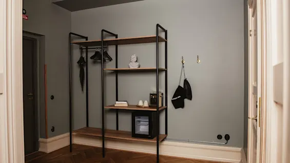 A shelving unit holds a bust, books, cups, a small fridge, and hangers in a minimalist room with wooden floors. A bag labeled "ANDOTHER" hangs on wall hooks.