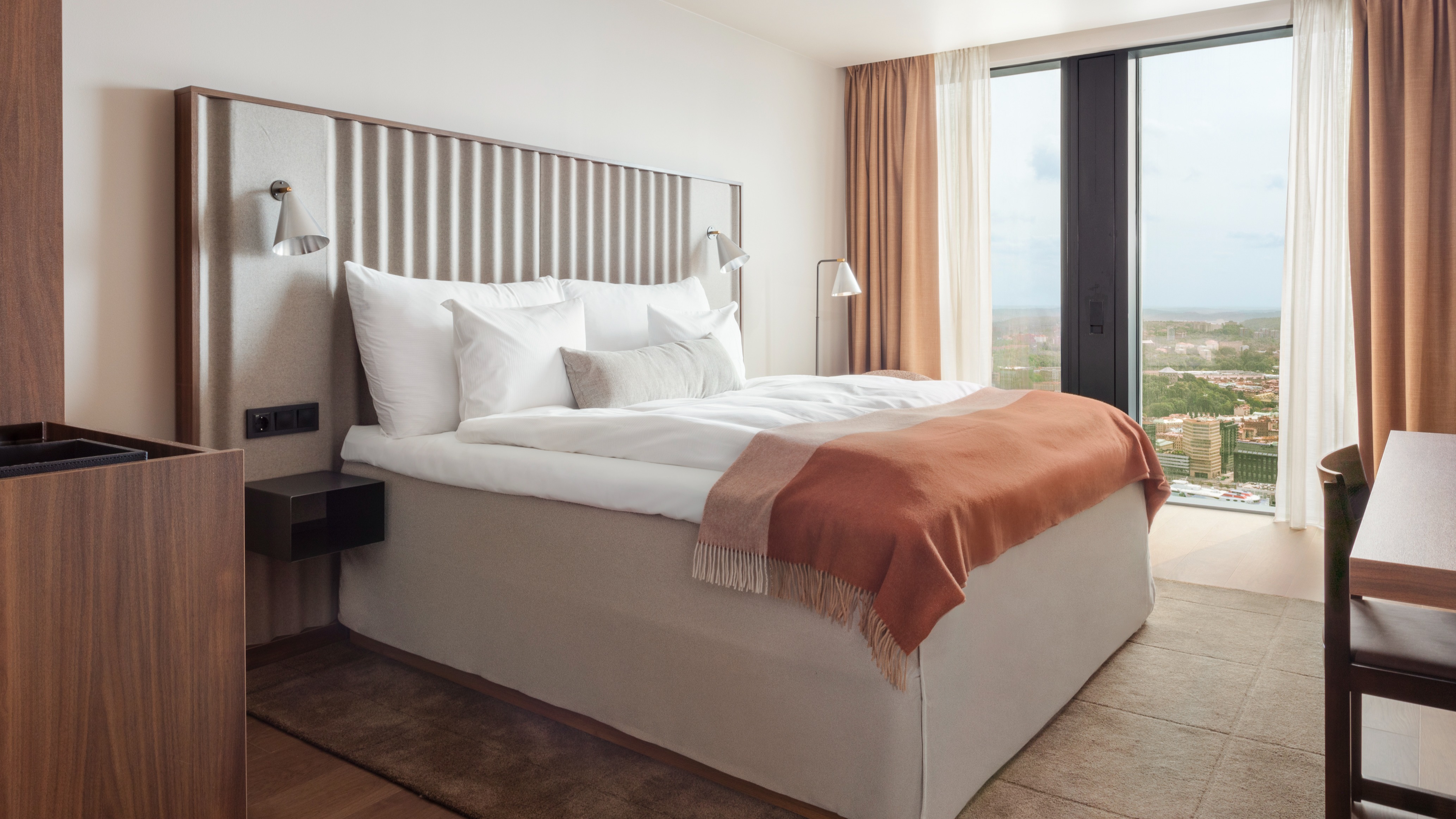 En seng med hvitt sengetøy og et oransje teppe står på et moderne hotellrom med beige gardiner og et stort vindu med utsikt over byens landskap.
