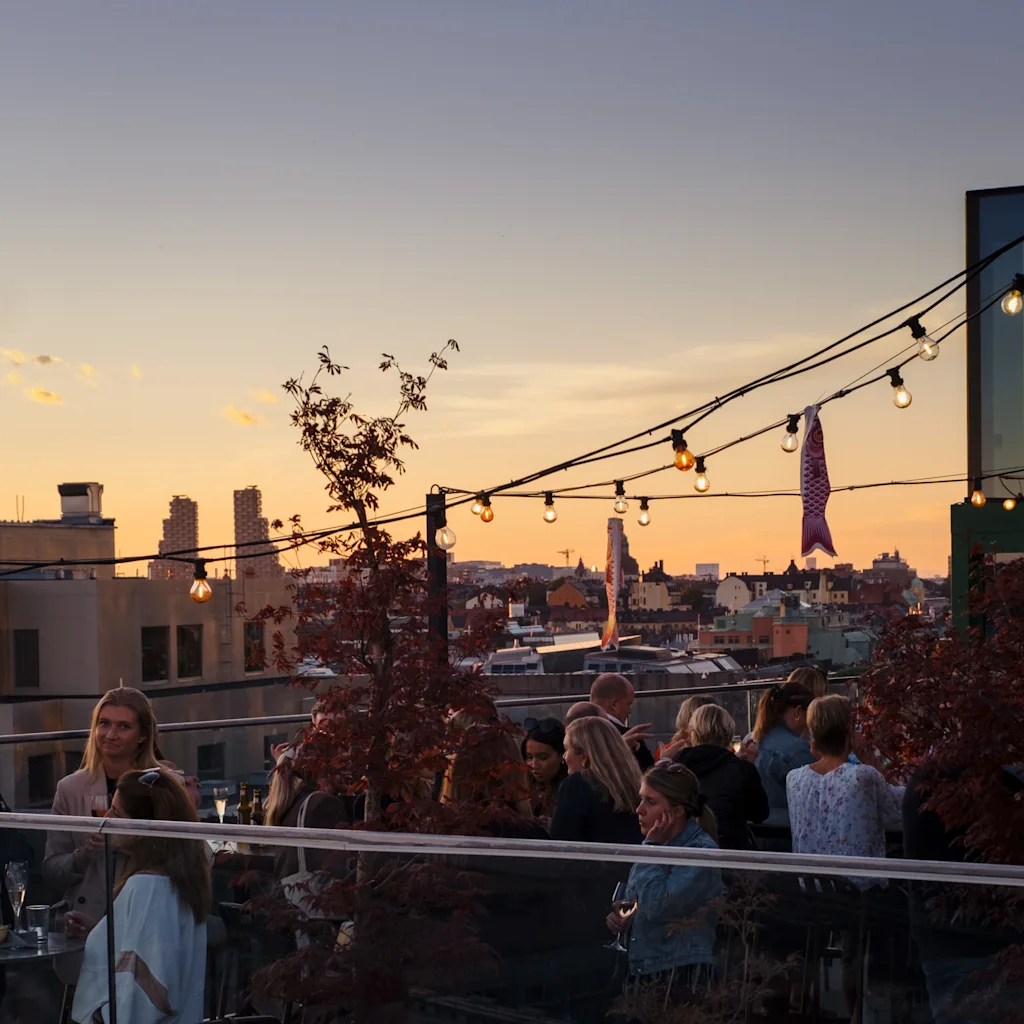 Gæster nyder drinks og samtaler på en livlig tagbar ved solnedgang, med udsigt over byen. TAK på Hobo i Stockholm.