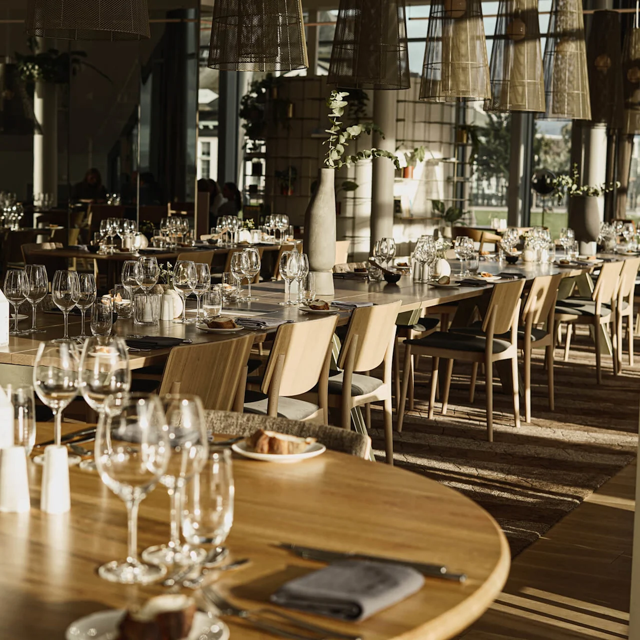 Et spisebord med pent arrangerte vinglass og bestikk, omgitt av trestoler. Mykt sollys filtrerer gjennom store vinduer, og gir en varm og innbydende atmosfære til restauranten.