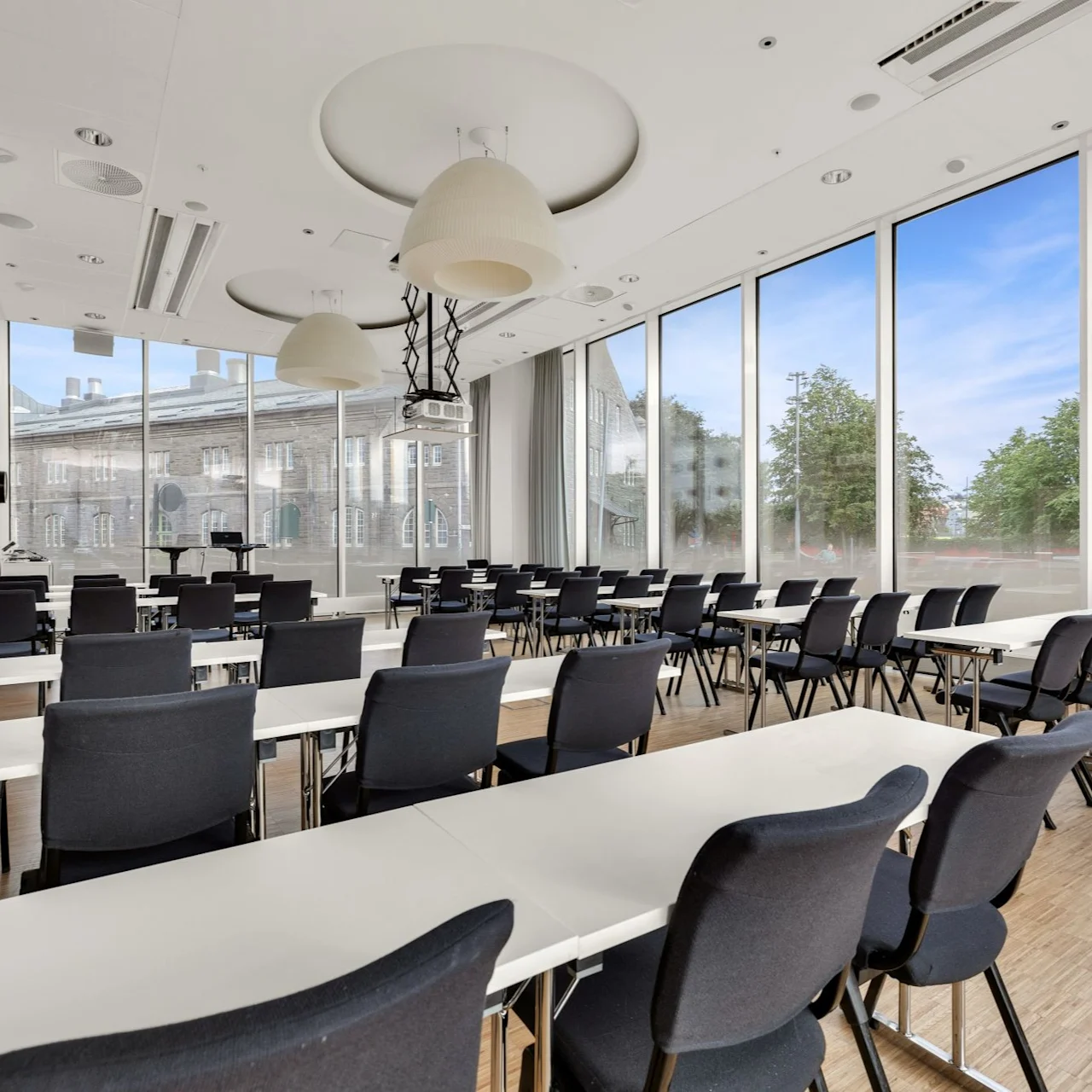 Classroom with black chairs and white tables, facing large windows. Projector and ceiling lights hang above. Trees and buildings visible outside under a blue sky.