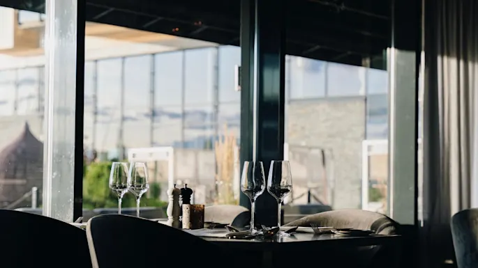 A set dining table with empty wine glasses is positioned indoors near large windows, overlooking a sunlit outdoor patio area.