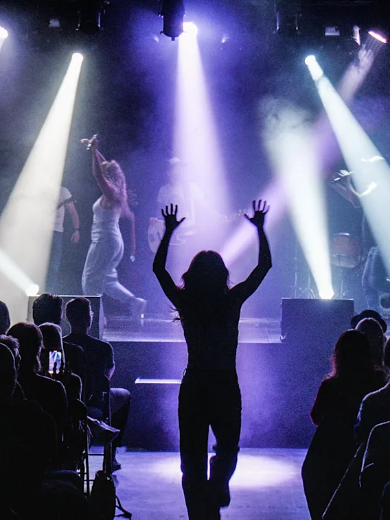 En livlig konsertscene med publikum som ser artister på scenen under lilla og hvite scenelys. En person i publikum har hendene hevet.
