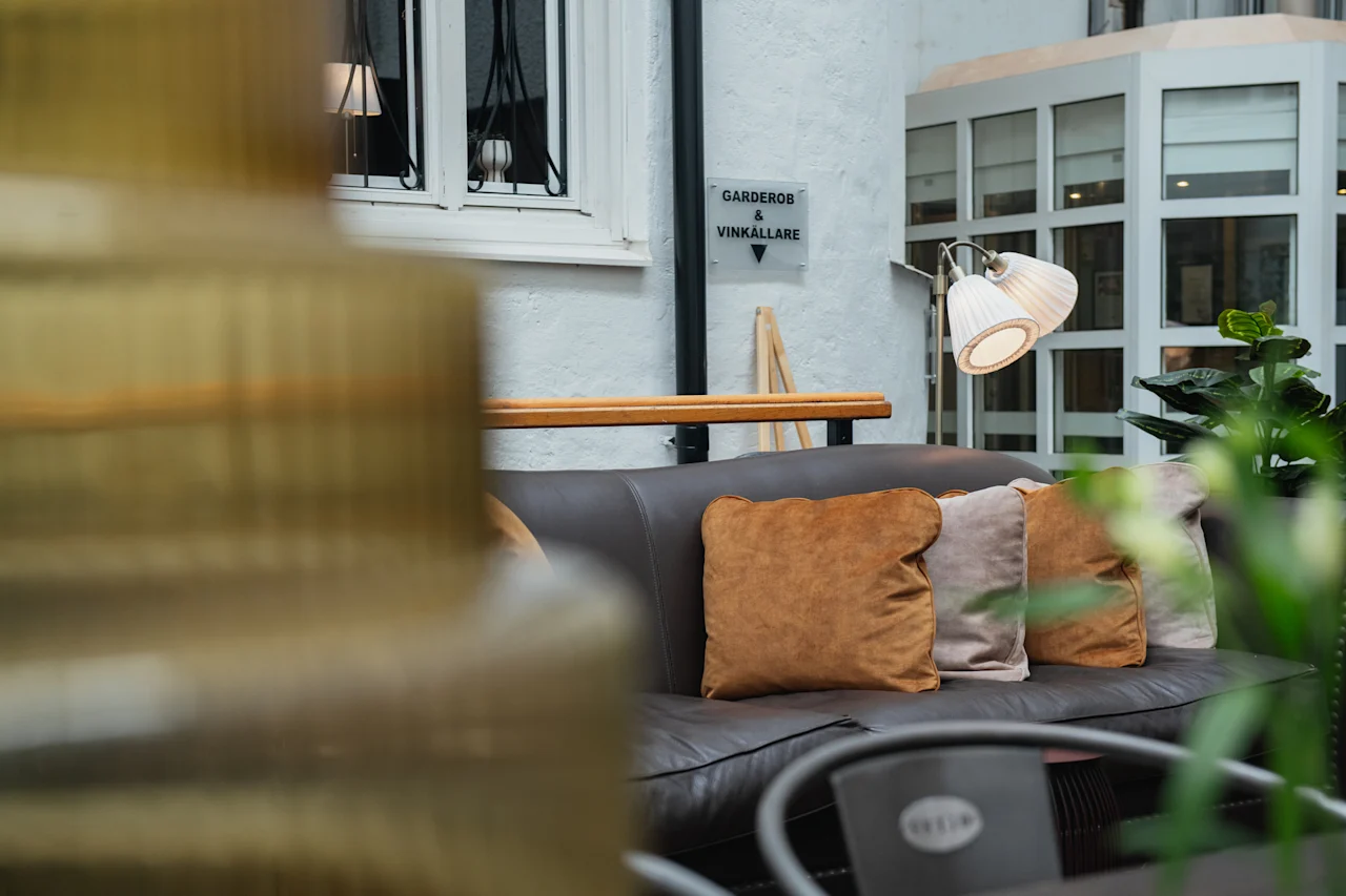 En soffa med beige kuddar står mot en vit vägg. En skylt lyder "Garderobe Vinkällare." En lampa lyser upp rummet, med växter som tillför grönska.