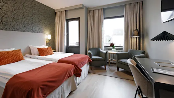Ett välupplyst hotellrum med två sängar, eleganta möbler, ett skrivbord och utsikt över staden från fönstren.