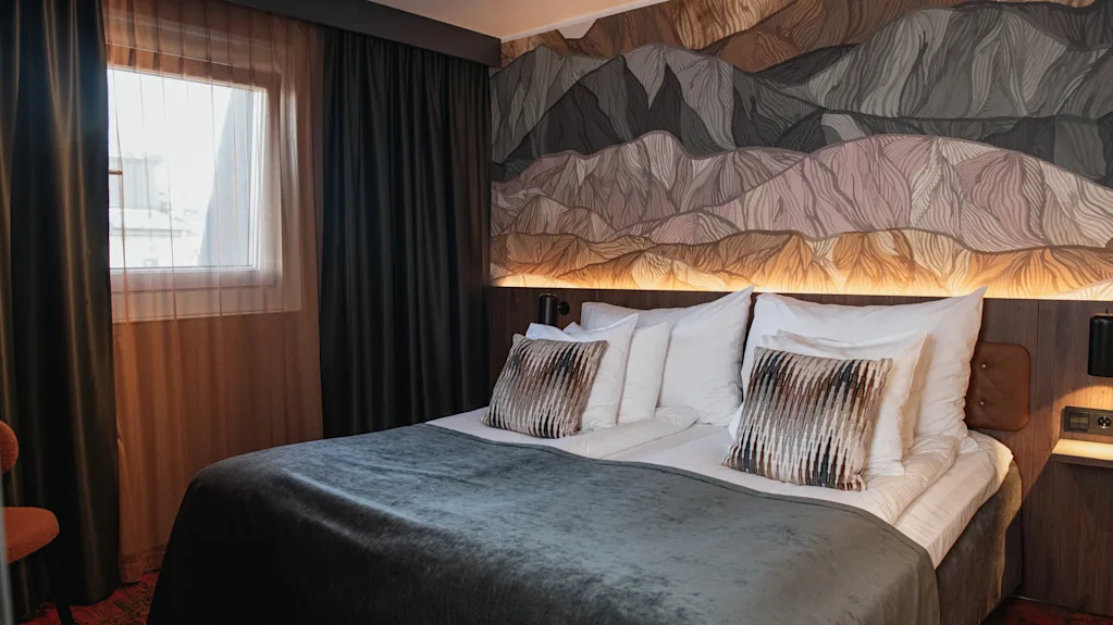 Et koselig hotellrom med en stor seng med grått teppe, et fjellinspirert veggmaleri og et vindu med gardiner.