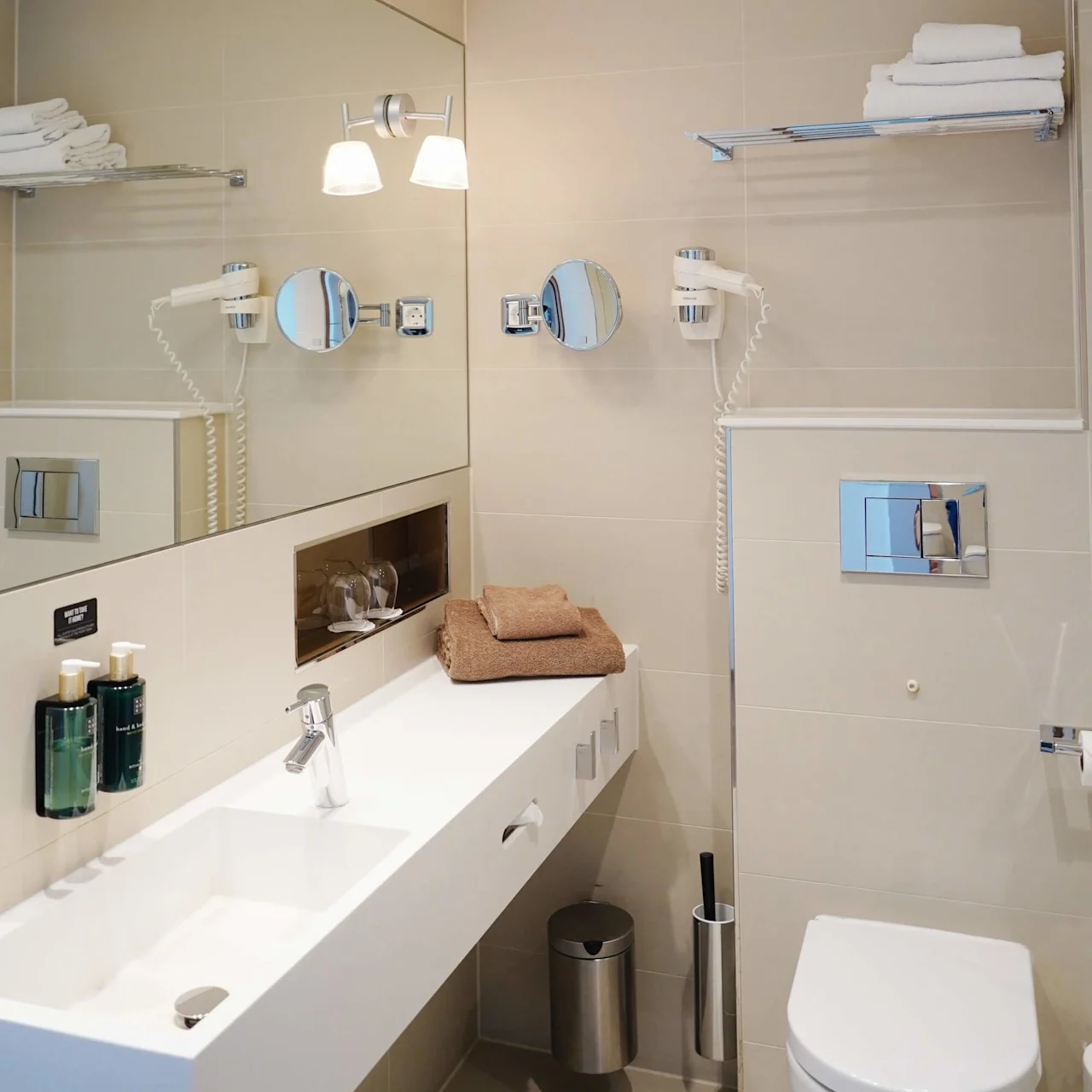 A modern bathroom features a white sink on a beige countertop with folded towels, under a large mirror. Wall-mounted toiletries and a hairdryer frame the space. Text: "HAND SOAP" and "BODY LOTION".