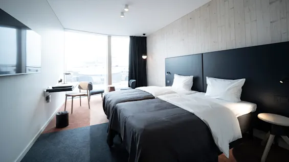 A modern hotel room features two neatly made beds with white linens, facing a wall-mounted television. A large window provides city views, complemented by minimalist furniture and soft lighting.