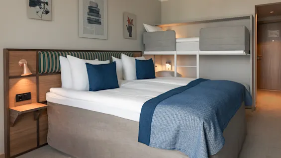 Et koselig hotellrom med en stor seng og blå detaljer, pluss en innebygd køyeseng, ideelt for familier eller grupper.