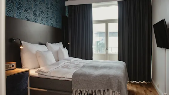 A neatly made bed with a gray blanket and white pillows is situated in a small room, flanked by dark curtains and wallpaper, lit by wall lamps, with a window view.
