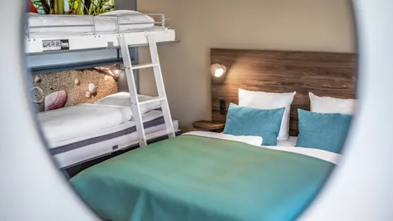 A double bed with teal bedding and two white pillows sits next to a bunk bed and ladder in a cozy modern bedroom with wooden accents. 