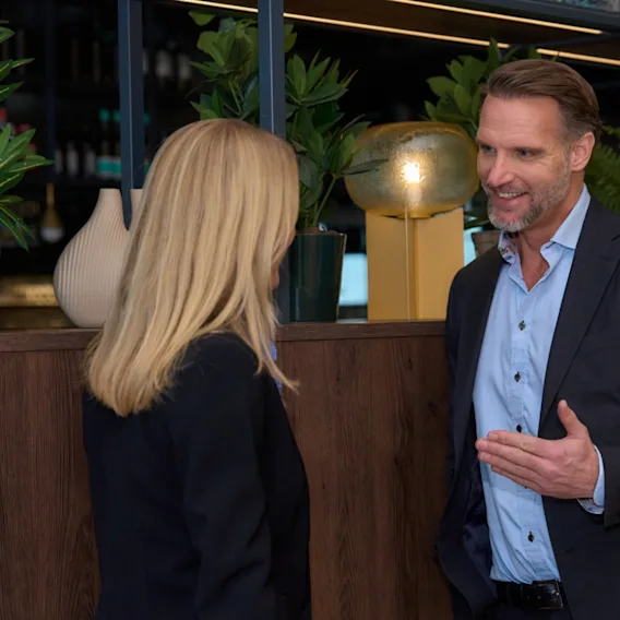 A man in a suit gestures while conversing with a woman in a business setting, surrounded by potted plants on a wooden counter.