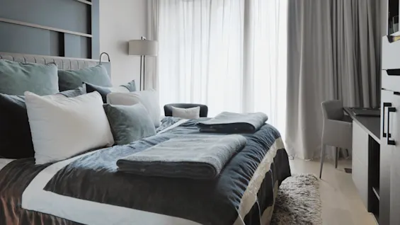 En pent, oppredd seng med lagdelte puter og brettede tepper står ved siden av høye vinduer med gjennomsiktige gardiner i et moderne soverom, med en gulvlampe og et skrivebord med stol.
