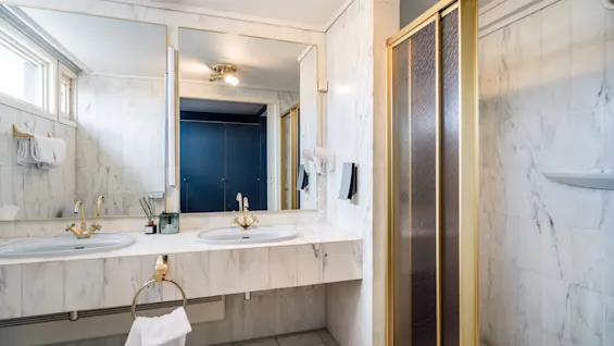 Et luksuriøst bad med marmorvegger, doble vasker, gullfargede armaturer og en dusj.