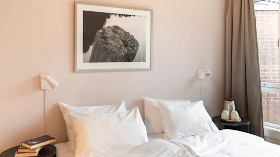 Et koselig soverom med en pent redd seng, nattbord med bøker og en vannkoker, samt et innrammet bilde.