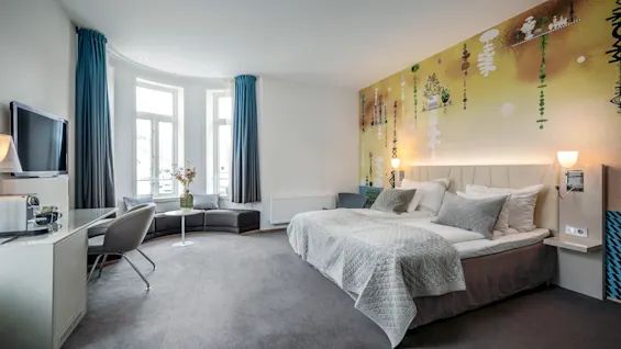 En stor säng med grå sängkläder finns i ett rymligt, modernt hotellrum. En färgglad, konstnärlig väggmålning pryder bakgrunden. En soffa, fönster, TV och skrivbord fullbordar den mysiga miljön.