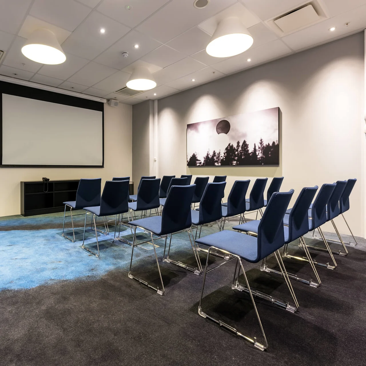 Rows of blue chairs face a large blank projection screen. The room has a gray carpet, blue accents, and artwork on the walls, illuminated by ceiling lights.