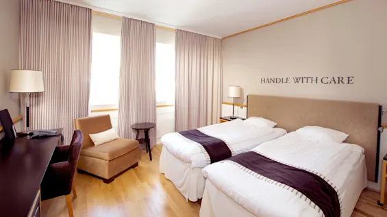Två enkelsängar med mörka accenter i ett upplyst hotellrum, randiga gardiner, möbler och väggkonst som säger "HANDLE WITH CARE."