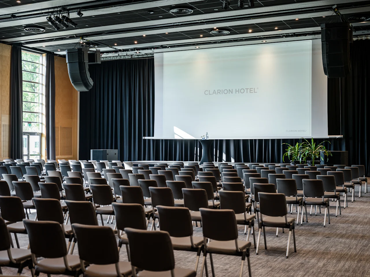 Ett rymligt konferensrum med rader av stolar vända mot en stor projektionsduk, redo för en presentation eller ett evenemang.