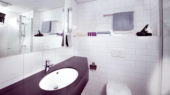 Et moderne badeværelse med hvide flisebelagte vægge, et glasdusch og toiletartikler.