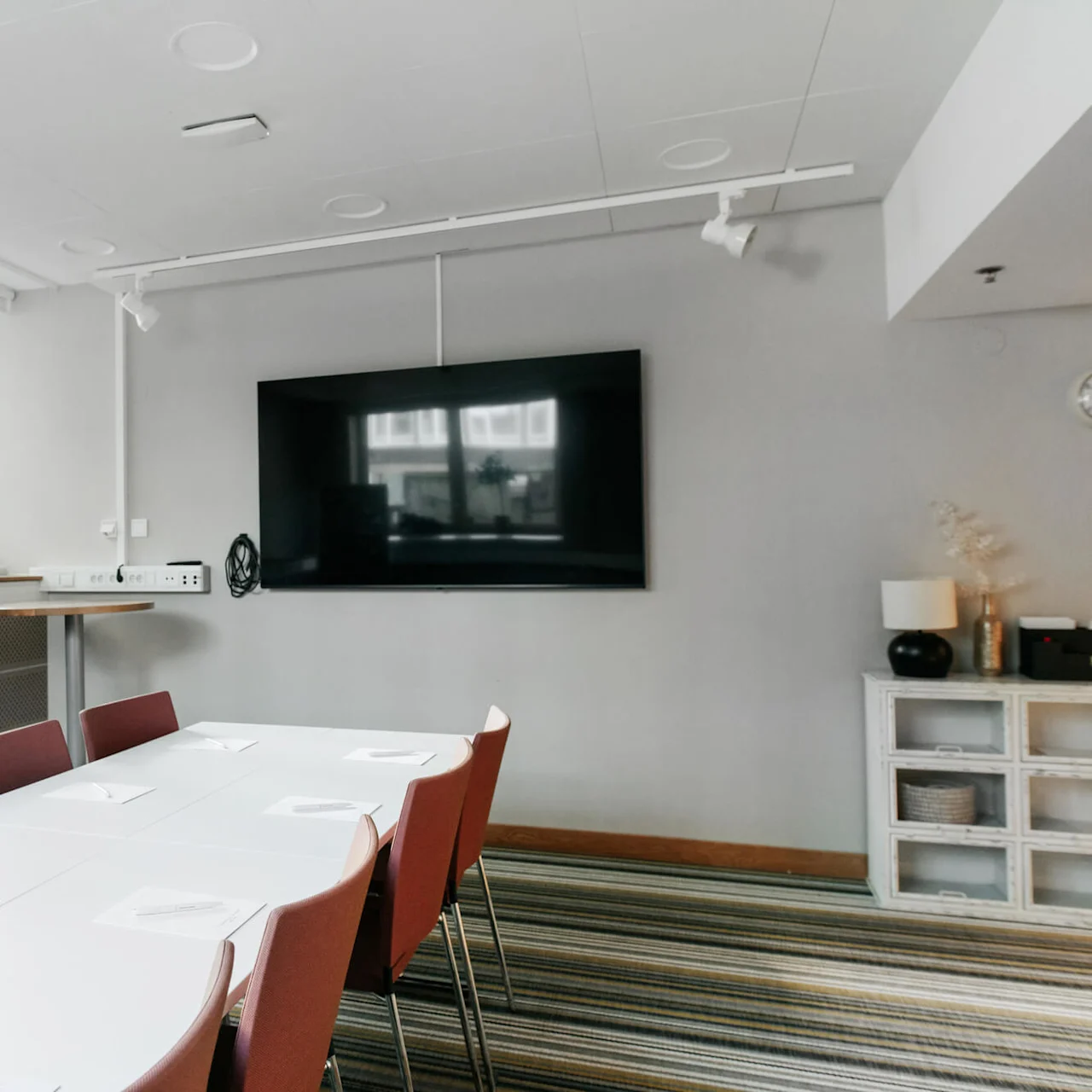 A modern meeting room with a long white table, chairs, and a large TV screen on the wall.