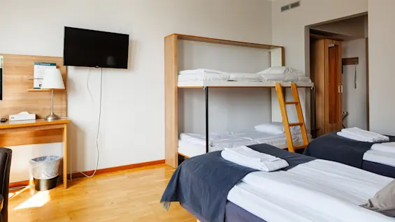 Våningssängar med en trästege står bredvid en prydligt bäddad säng i ett välstädat hotellrum. En väggmonterad TV vetter mot ett skrivbord med lampa och näsdukshållare. 