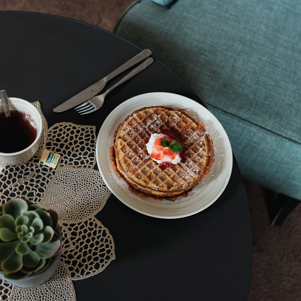 A plate with waffles topped with whipped cream and strawberries on a table. Beside it, a cup of tea with a teabag and a succulent plant sit on a woven placemat.