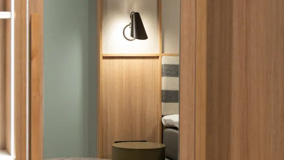 Et hyggeligt hotelværelseinteriør med seng, moderne lampe og sideborde, med lysegrønne vægge og trædetaljer.