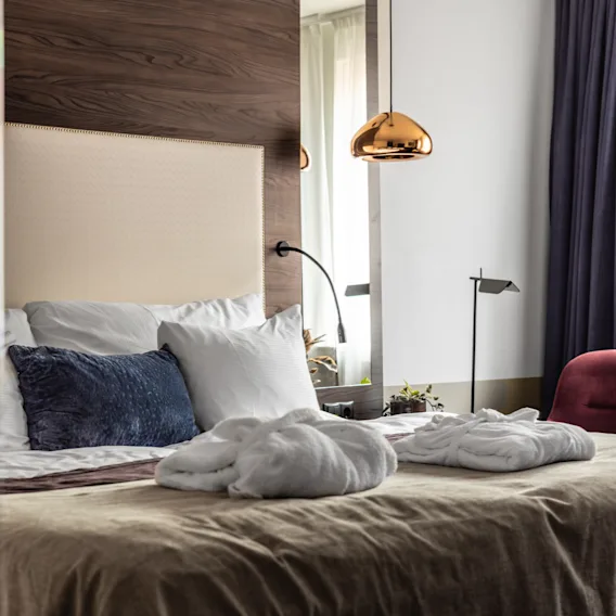 Ett mysigt hotellrum med en prydligt bäddad säng, dekorerad med kuddar och ett par morgonrockar. Mjukt ljus sipprar genom gardiner bredvid en stilfull röd stol. En varm och inbjudande atmosfär fyller rummet.